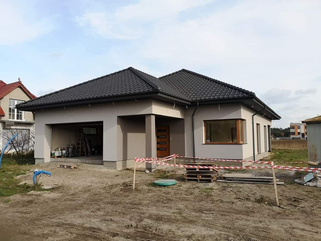 Nowoczesny, parterowy dom z szarą elewacją i grafitową dachówką, częściowo wykończony, z otwartym garażem, widoczne ślady prac budowlanych w otoczeniu.