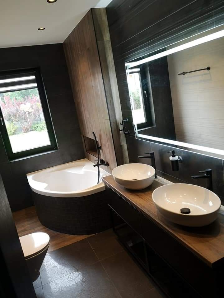Nowoczesna łazienka z wanną narożną obudowaną ciemnymi płytkami, dwiema okrągłymi umywalkami nablatowymi i podświetlanym lustrem.