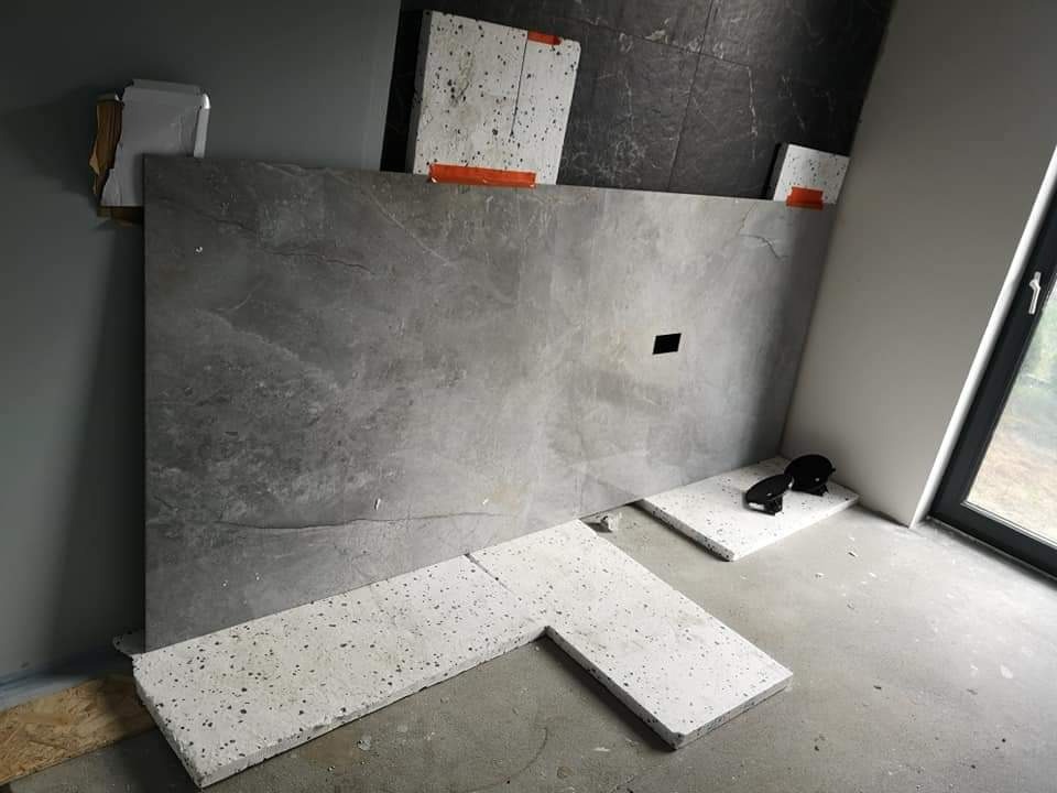 Montaż dużej płyty z szarego kamienia z czarnym otworem na ścianie w łazience, z fragmentami białych płytek z czarnymi plamkami na podłodze, widoczne narzędzia i okno.