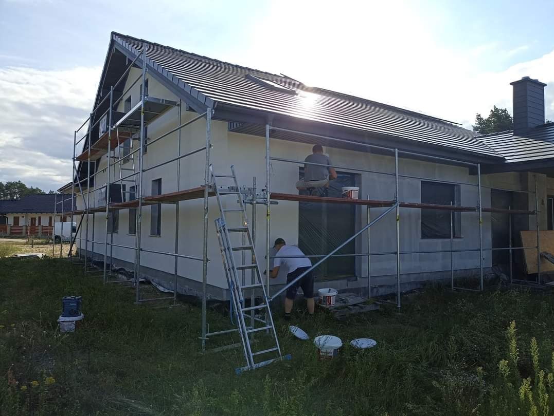 Dwóch pracowników na rusztowaniu przy dwukondygnacyjnym domu z ciemnym dachem, jeden maluje ścianę, drugi siedzi na rusztowaniu, widoczne wiaderka z farbą i drabina.