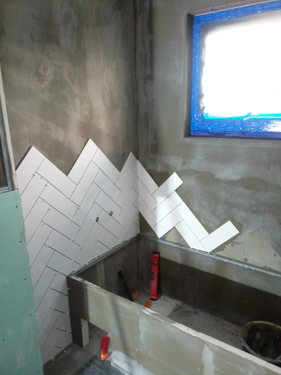 Układanie białych, prostokątnych płytek w jodełkę na ścianie w surowej łazience, widoczna wanna w trakcie obudowy, poziomica i okno zabezpieczone folią.