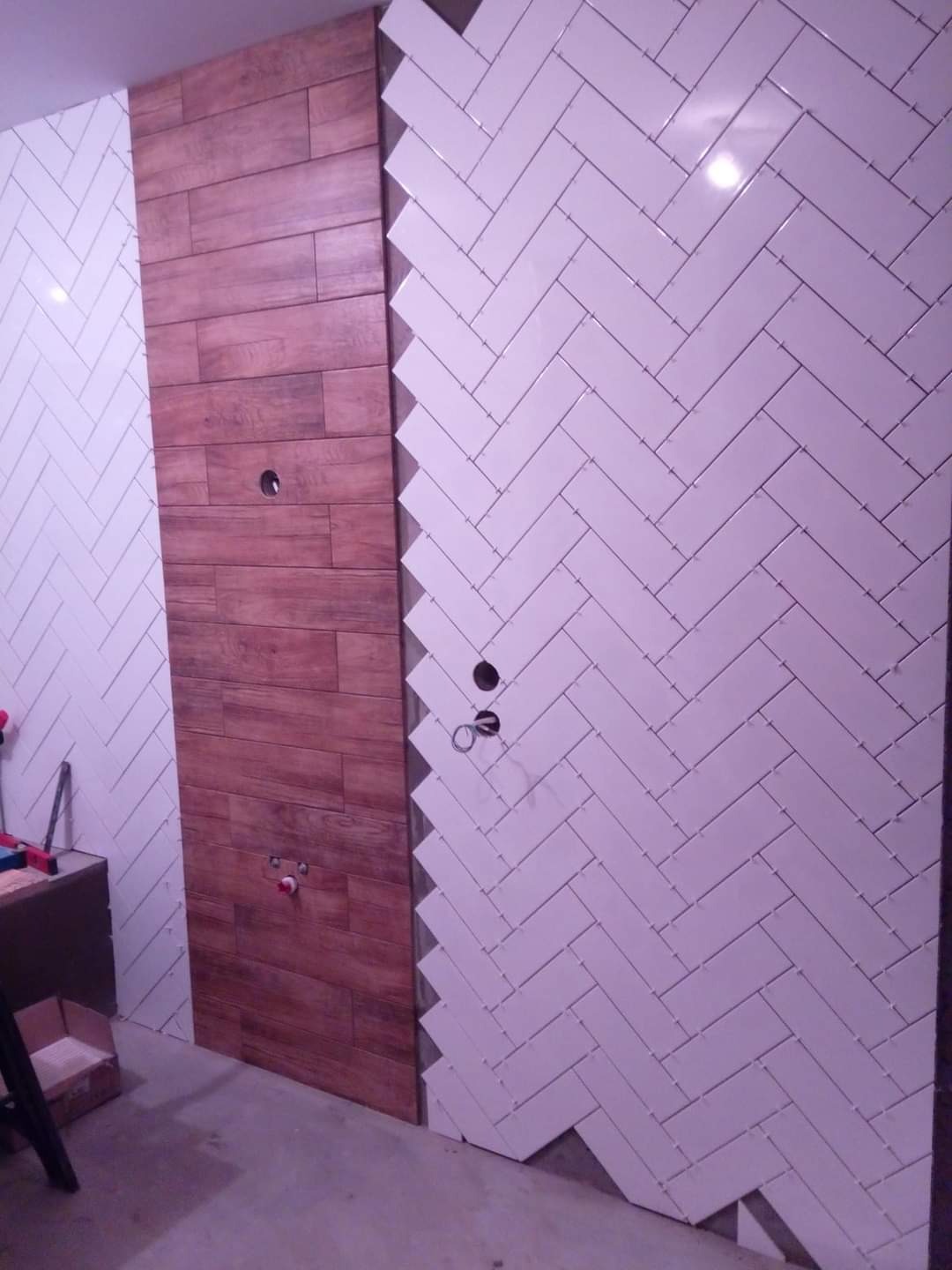 Ściana w trakcie wykańczania z dwoma rodzajami płytek: drewnopodobne ułożone poziomo oraz białe, małe, ułożone w jodełkę, z widocznymi otworami na instalację hydrauliczną.