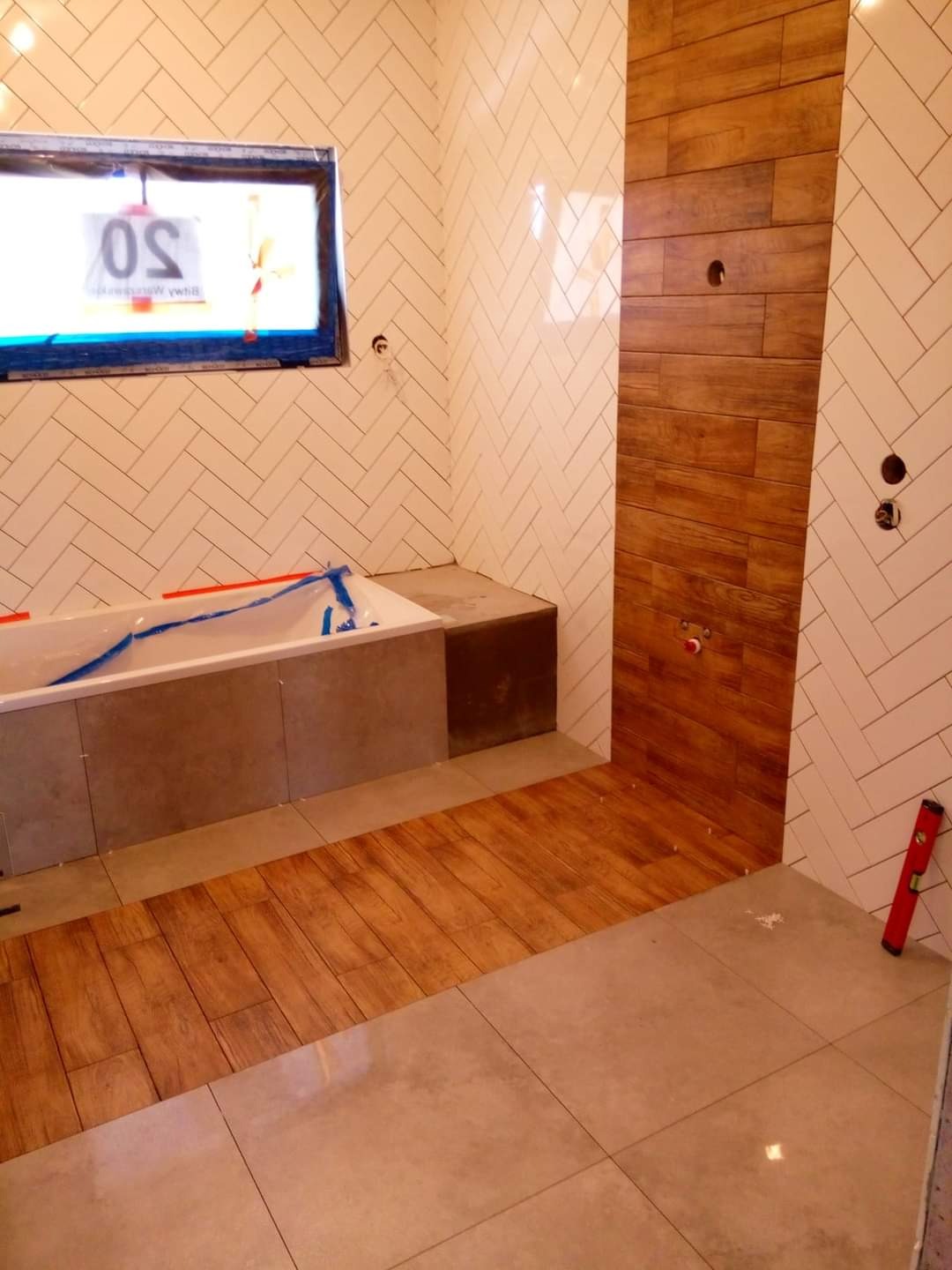 Wnętrze łazienki w trakcie prac wykończeniowych: ułożone płytki na ścianach w wzór jodełki, drewnopodobna okładzina ściany, częściowo ułożona podłoga z płytek drewnopodobnych i szarych, wanna...