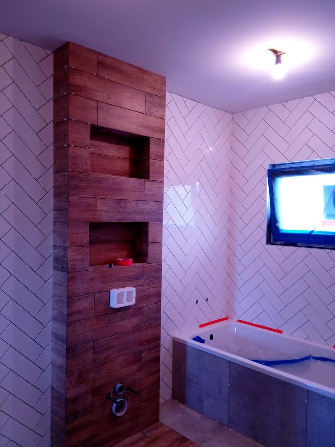 Wnęka z drewnopodobnych płytek z dwoma półkami w łazience z białymi płytkami ułożonymi w jodełkę, widoczne przyłącza hydrauliczne i zabezpieczona wanna.