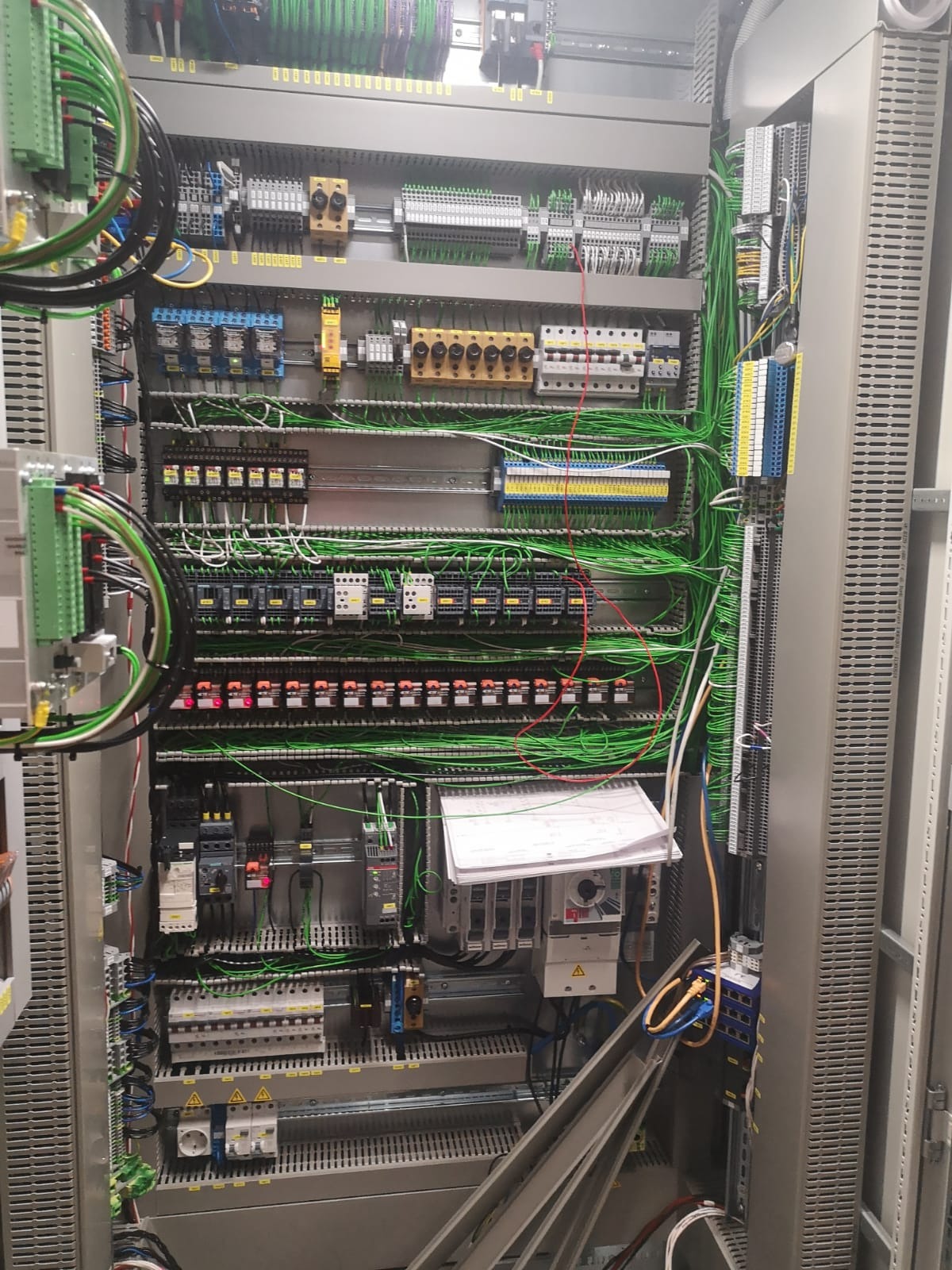 Otwarta szafa sterownicza z gęstą siecią przewodów, listwami zaciskowymi i modułami sterującymi, z dokumentacją techniczną leżącą na dolnej części szafy.