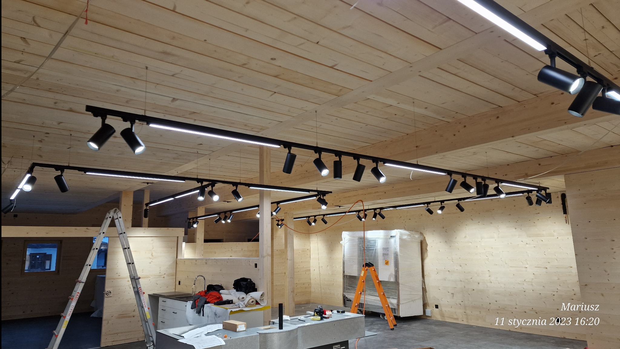 Montaż oświetlenia szynowego z reflektorami w pomieszczeniu wykończonym drewnem, widoczne drabiny i narzędzia.