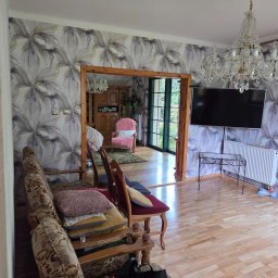 Kornobud - Salon z wzorzystą tapetą w kwiaty, drewnianą podłogą i kryształowym żyrandolem. Widoczne meble w stylu retro i otwarte przejście do innego pomieszczenia.