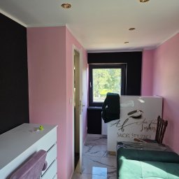 Kornobud - Jasny pokój z różowymi ścianami i czarnym akcentem. Widoczne białe meble, okno i lustrzana podłoga. Wnętrze po malowaniu, gotowe do użytku. Salon stylizacji rzęs.