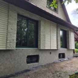 Elewacja domu z sidingiem, kamiennymi wstawkami wokół okien i płytkami na fundamencie. Widoczne okna z ciemnymi ramami i fragment dachu.