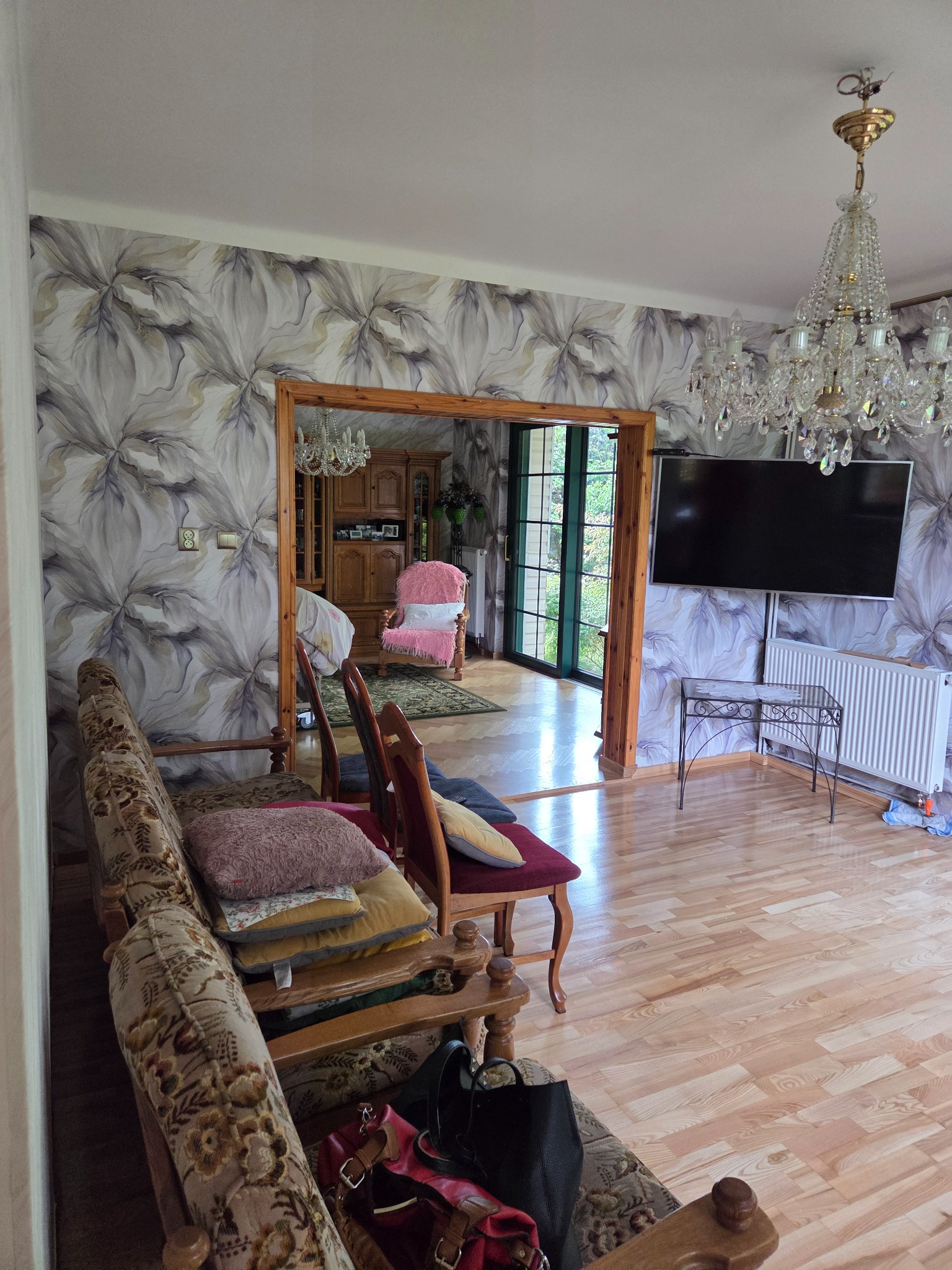 Salon z wzorzystą tapetą w kwiaty, drewnianą podłogą i kryształowym żyrandolem. Widoczne meble w stylu retro i otwarte przejście do innego pomieszczenia.