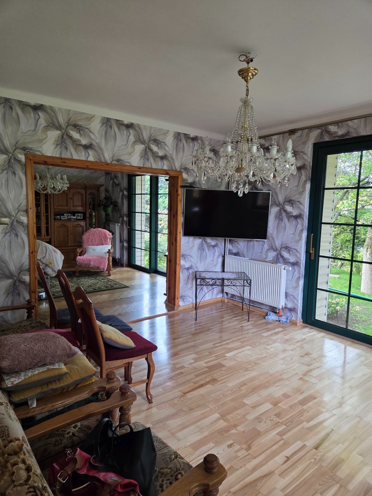 Eleganckie wnętrze: kwiatowe tapety, kryształowy żyrandol i parkiet. Widok na pokój z różowym fotelem przez drewniane obramowanie. Duże okna wpuszczają światło.