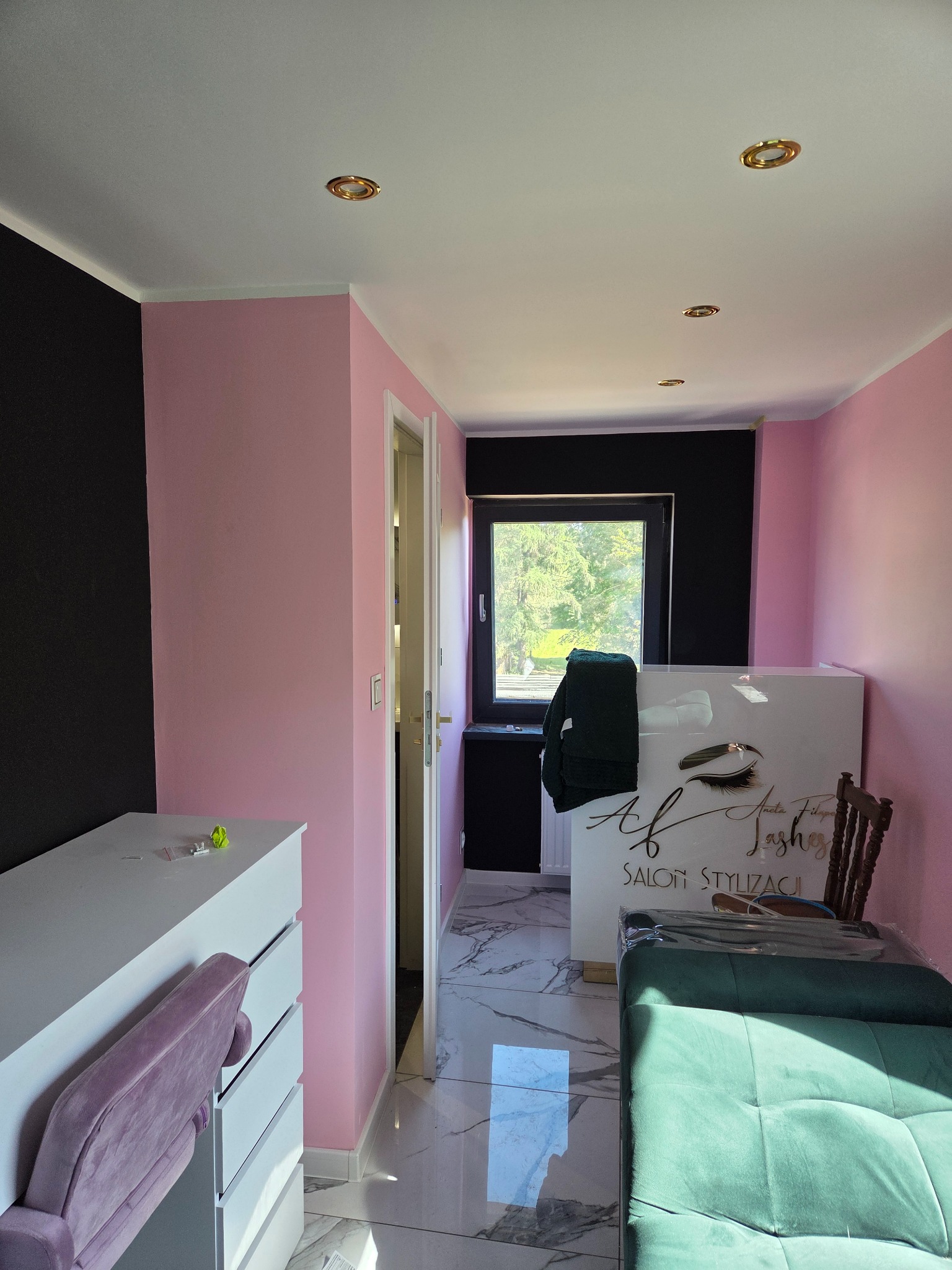 Jasny pokój z różowymi ścianami i czarnym akcentem. Widoczne białe meble, okno i lustrzana podłoga. Wnętrze po malowaniu, gotowe do użytku. Salon stylizacji rzęs.