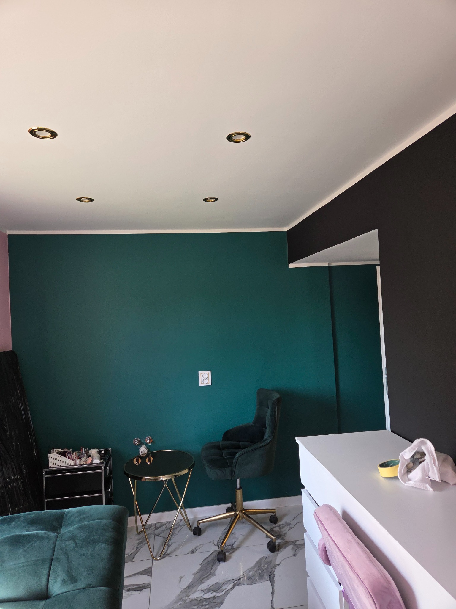 Pokój po malowaniu ścian na butelkową zieleń i czarno. Widoczne meble i marmurowa podłoga. Sufit biały z okrągłymi lampami.