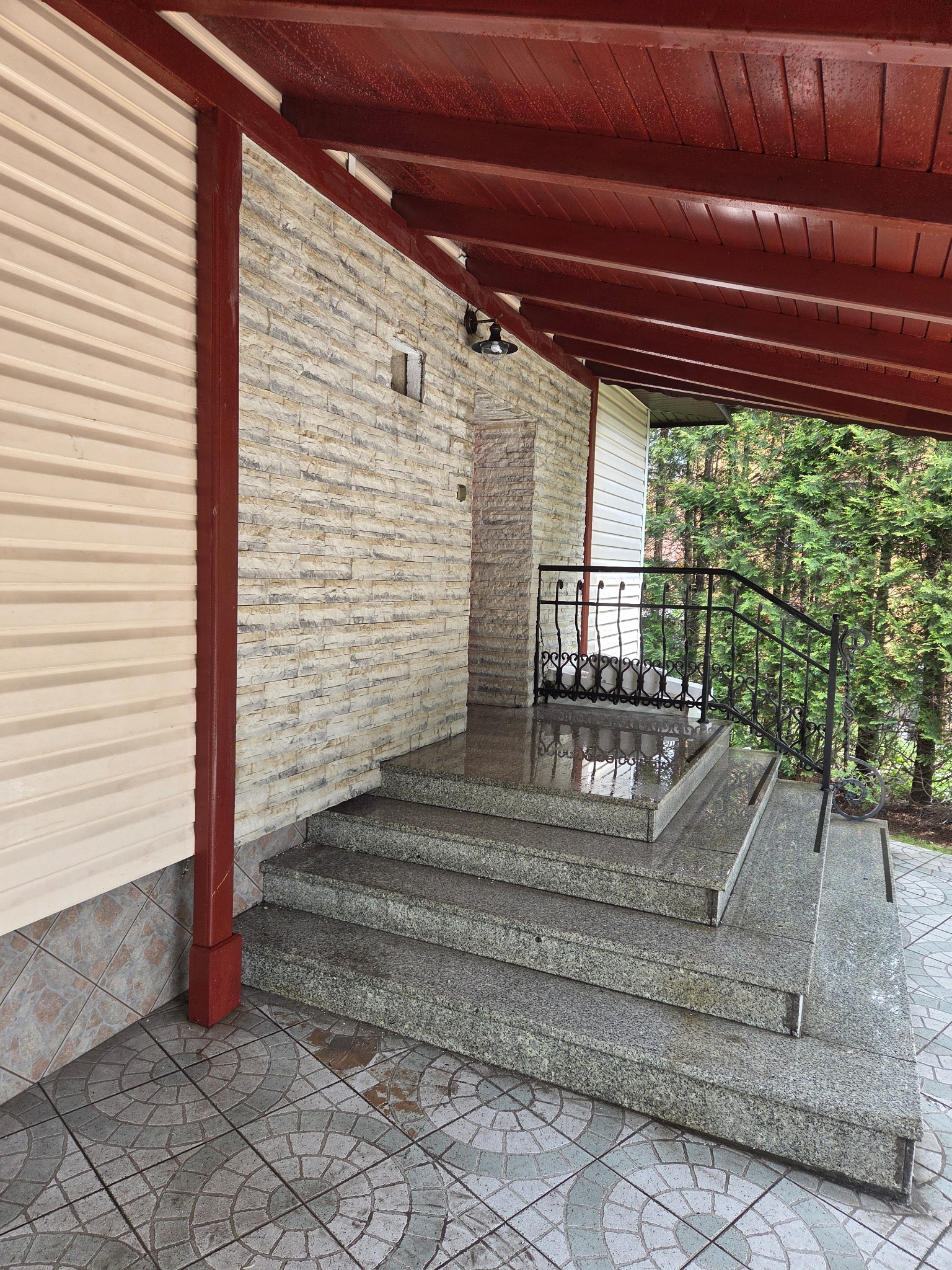 Zadaszone schody wejściowe z szarego granitu, z kutą balustradą. Kamienna elewacja budynku z kremową okładziną po lewej. Czerwona drewniana konstrukcja dachu.