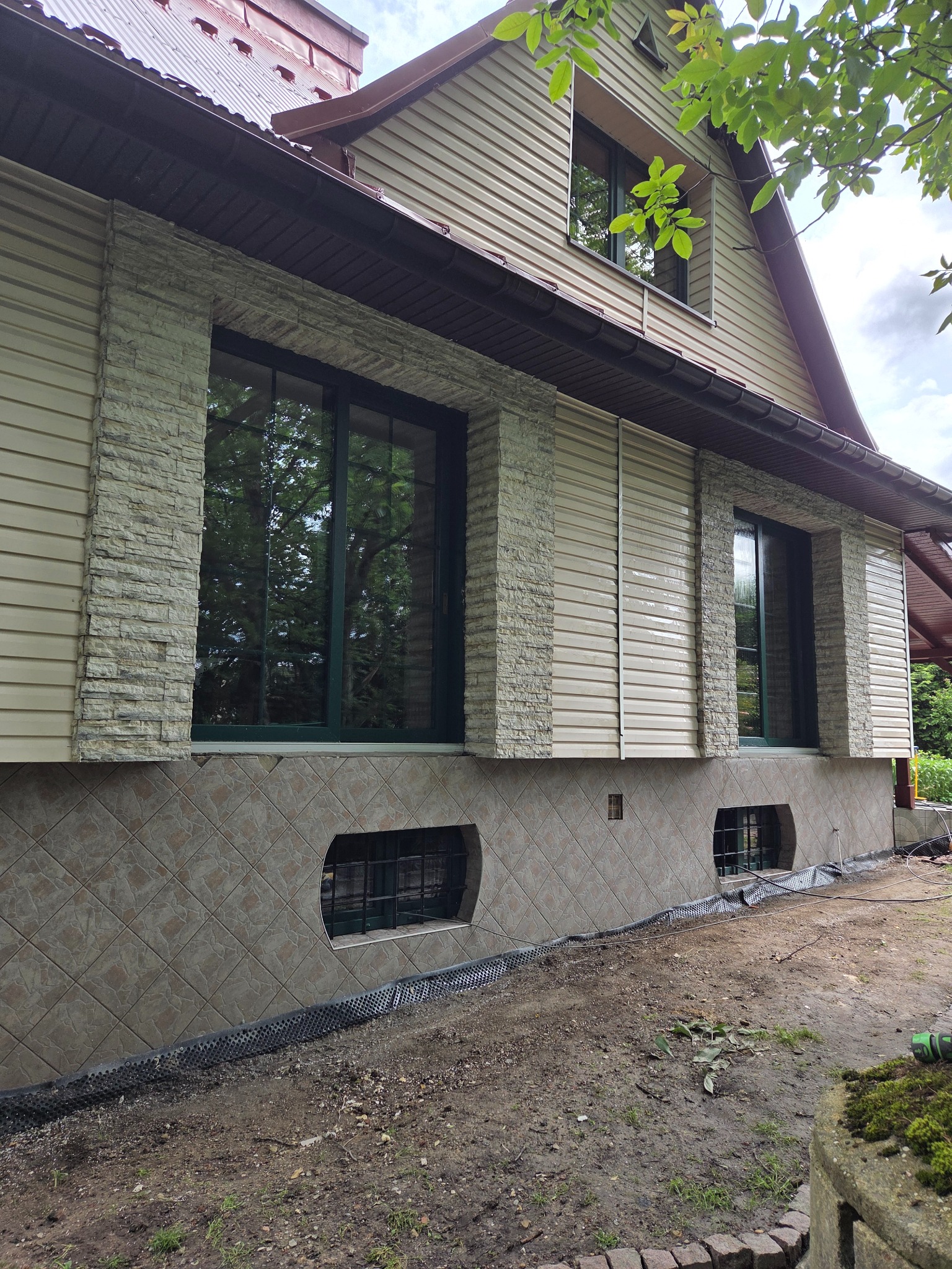 Elewacja domu z sidingiem, kamiennymi wstawkami wokół okien i płytkami na fundamencie. Widoczne okna z ciemnymi ramami i fragment dachu.