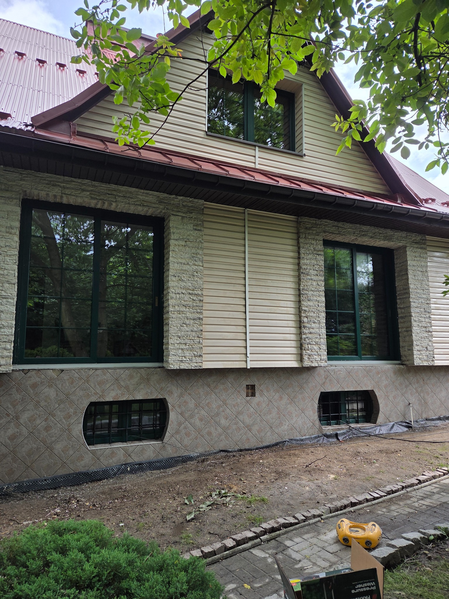 Dom z beżową elewacją sidingową, obramowany kamieniem i z ciemnozieloną stolarką okienną. Widoczne okna piwniczne i fragment ogrodu. Ujęcie z dołu.