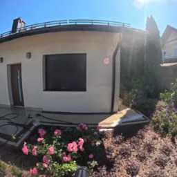 Jasna elewacja domu jednorodzinnego z czarnymi roletami i elementami dekoracyjnymi, otoczona zadbaną roślinnością i kostką brukową, widoczne węże do myjki ciśnieniowej.