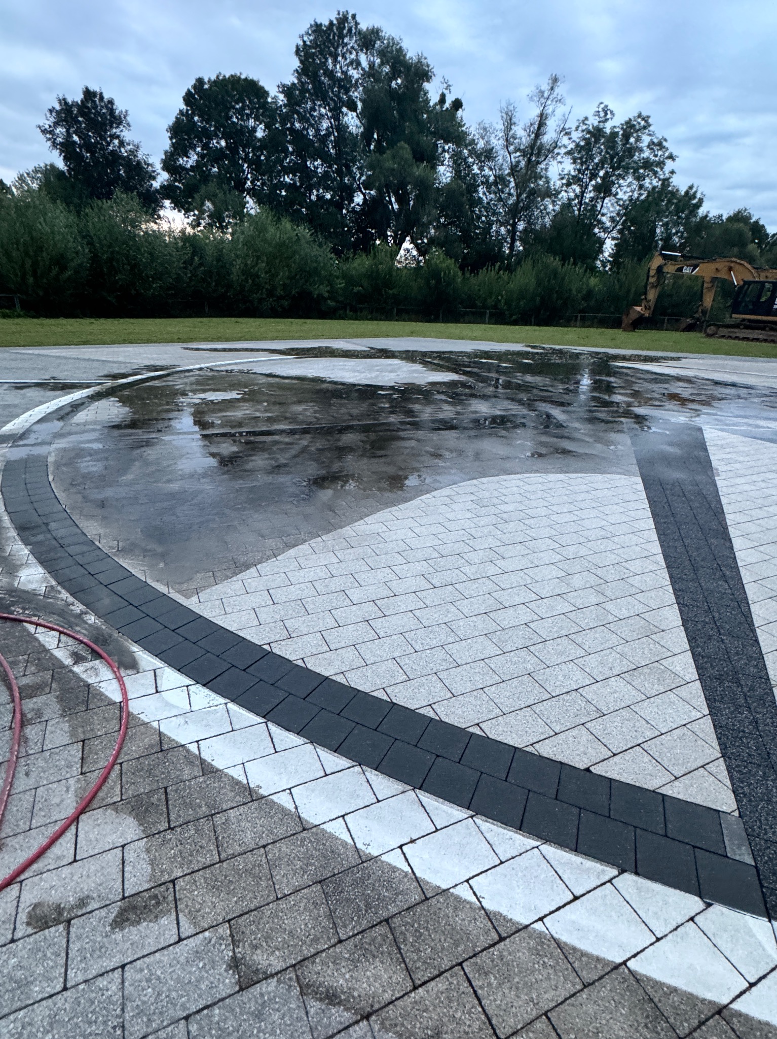 Mokra kostka brukowa po czyszczeniu, widoczny wąż ciśnieniowy i fragment koparki w tle.