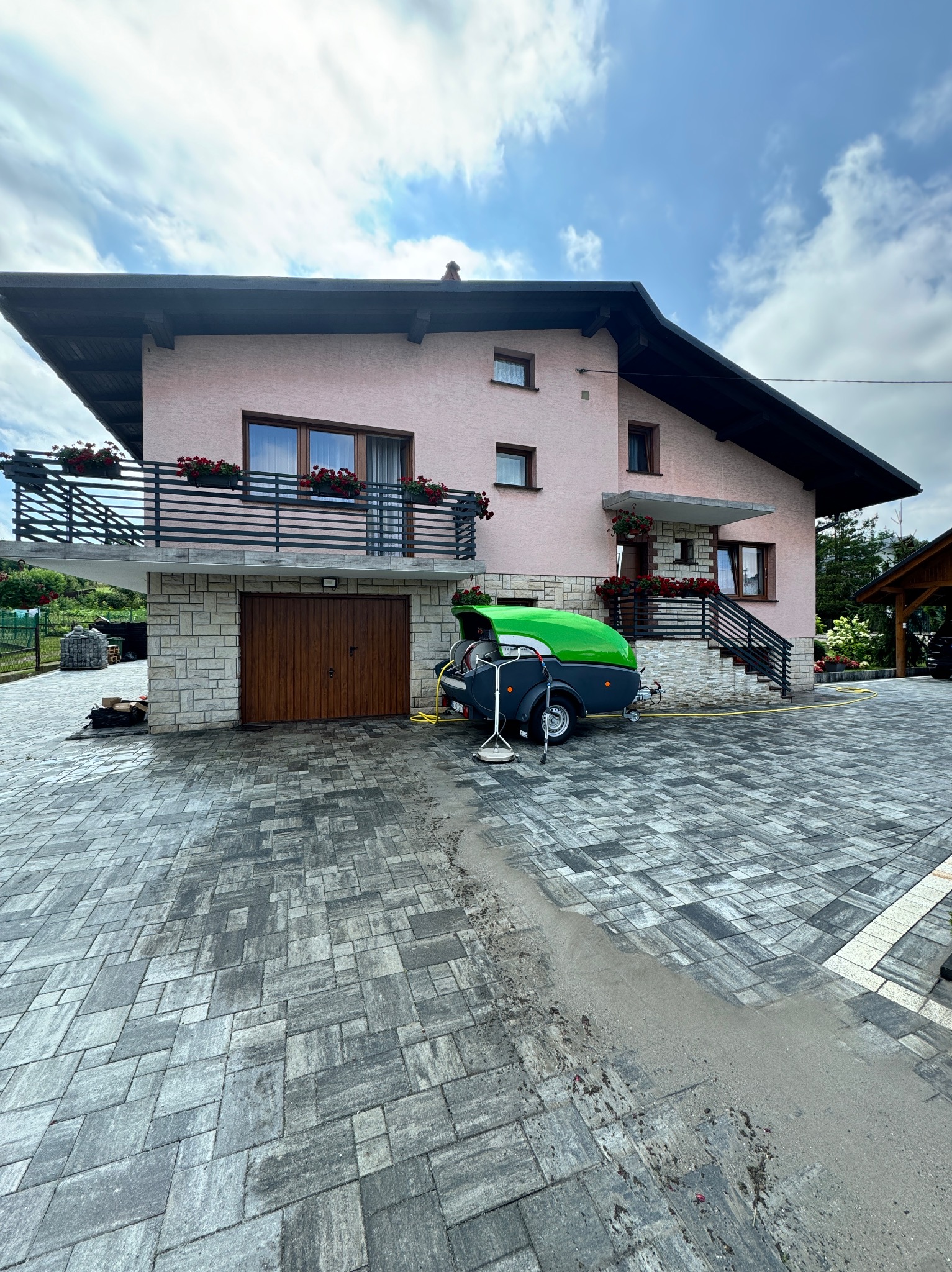 Dwupiętrowy dom z różową elewacją i szarą kostką brukową, z fragmentem wyczyszczonej powierzchni. Na podjeździe stoi przyczepa z urządzeniem do czyszczenia pod ciśnieniem.