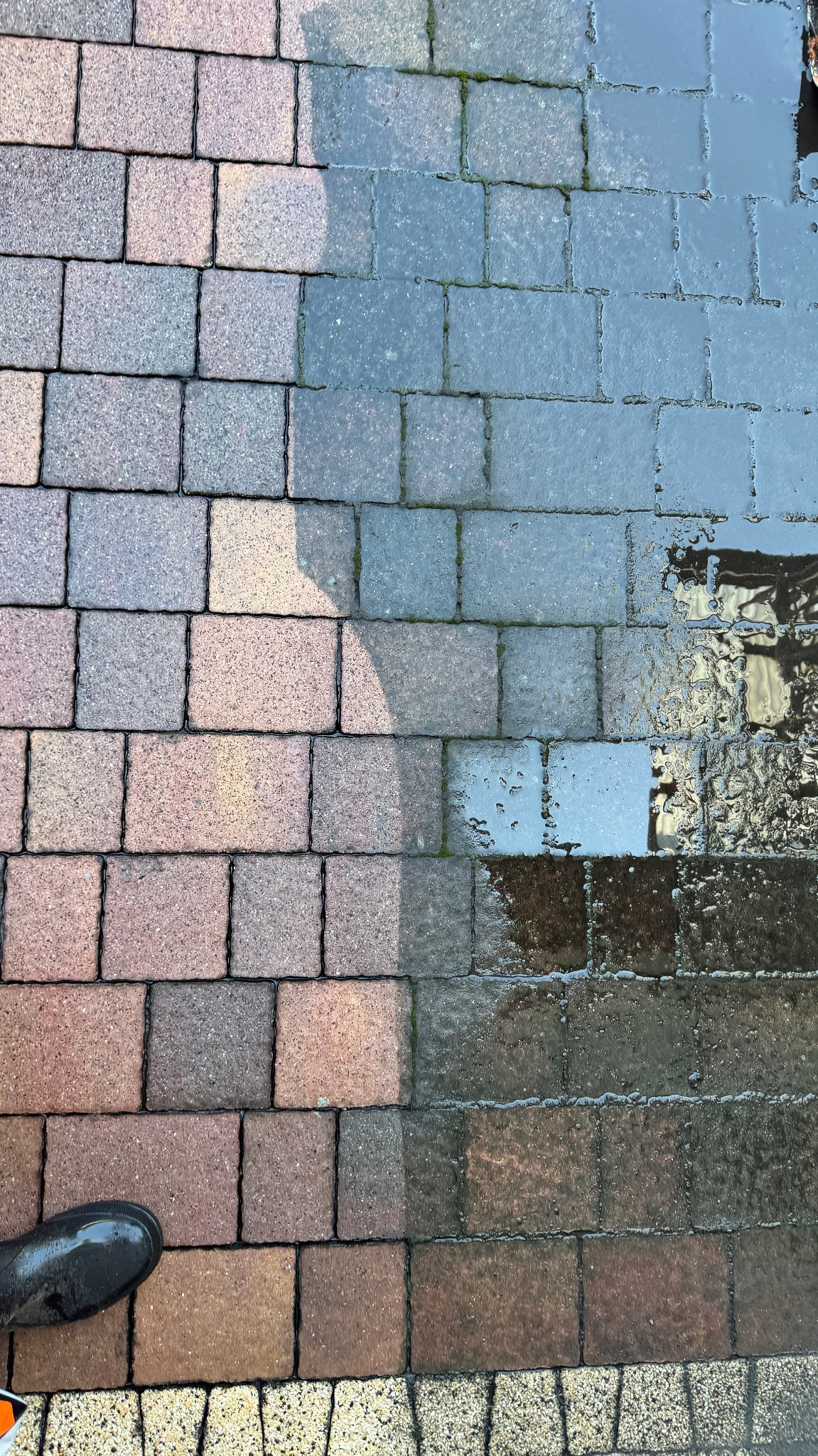 Częściowo wyczyszczona kostka brukowa z widoczną różnicą w kolorze i zabrudzeniach, gumowy but w rogu kadru.