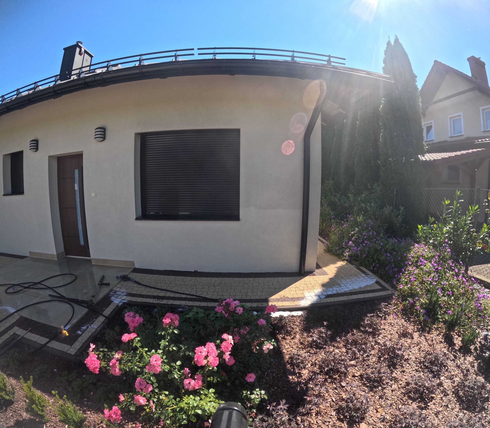 Jasna elewacja domu jednorodzinnego z czarnymi roletami i elementami dekoracyjnymi, otoczona zadbaną roślinnością i kostką brukową, widoczne węże do myjki ciśnieniowej.