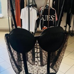 1LS Iryna Kozarenko - Czarny koronkowy gorset z tiulowymi wstawkami w kropki, zawieszony na wieszaku w butiku odzieżowym, w tle widoczne inne ubrania na wieszakach i regale.