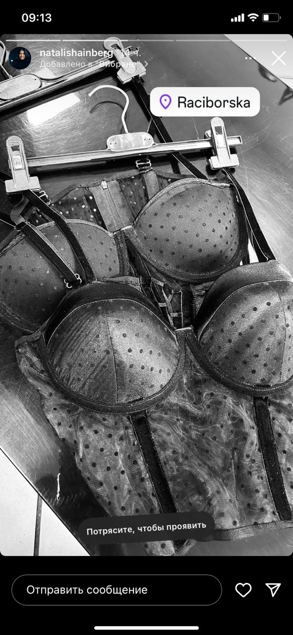 Czarno-białe zdjęcie biustonosza i gorsetu w drobne kropki, wiszących na wieszaku z metalowymi klipsami, widoczne fragmenty ekranu telefonu z tekstem w języku rosyjskim.