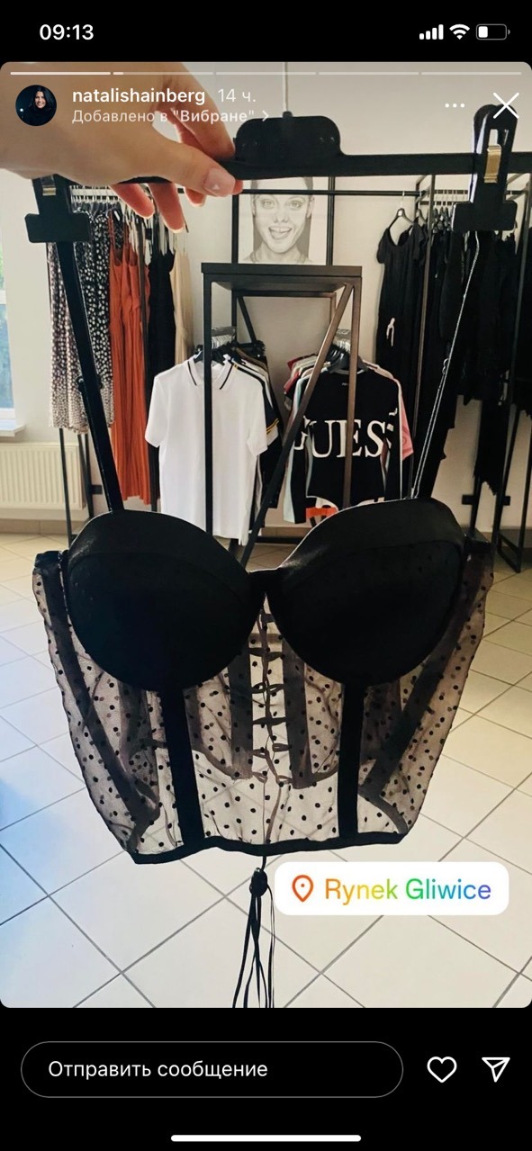 Czarny koronkowy gorset z tiulowymi wstawkami w kropki, zawieszony na wieszaku w butiku odzieżowym, w tle widoczne inne ubrania na wieszakach i regale.