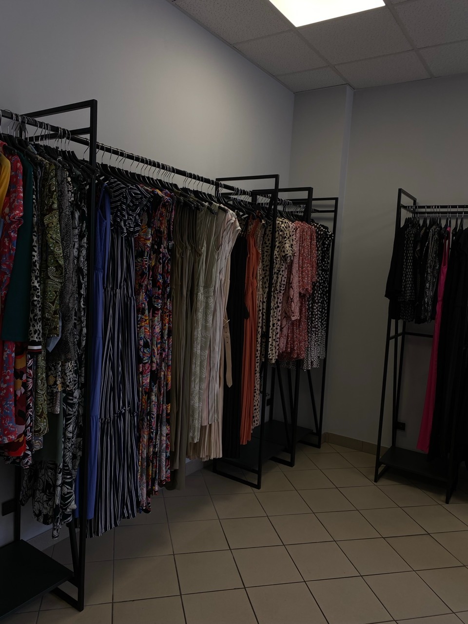 Rzędy wieszaków z różnorodnymi sukienkami w butiku, widoczne wzory kwiatowe, paski i plisy, oświetlenie górne, płytki podłogowe.
