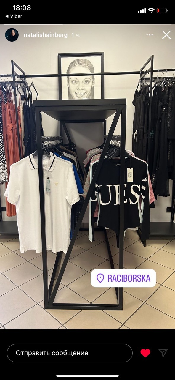 Wnętrze sklepu odzieżowego z czarnym metalowym regałem, na którym wiszą biała koszulka polo i czarny top z napisem GUESS, w tle wieszaki z różnymi ubraniami i portret kobiety w czarnej ramie.