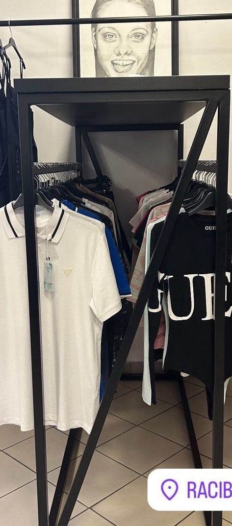 Wnętrze sklepu odzieżowego z czarnym metalowym regałem, na którym wiszą koszulki polo i t-shirty, nad regałem wisi oprawiony portret kobiety.