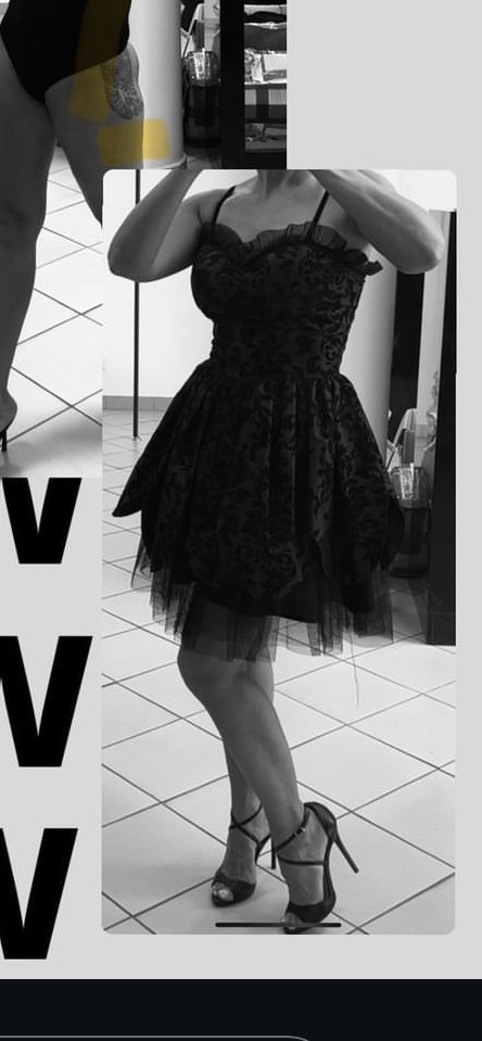 Czarno-białe zdjęcie kobiety w krótkiej, czarnej sukience z tiulowym dołem i butach na wysokim obcasie, pozującej na tle białej kafelkowej podłogi.