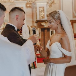 Myszyński Design Sp. z o. o. - Para młoda podczas ceremonii ślubnej w kościele, wymiana obrączek, panna młoda w białej sukni z odkrytymi ramionami i welonem, pan młody w ciemnym garniturze, ksiądz trzyma mikrofon.