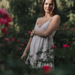 Portret młodej kobiety w letniej sukience z ramiączkami, pozującej w ogrodzie różanym, z delikatnym uśmiechem i brązową bransoletką na nadgarstku.