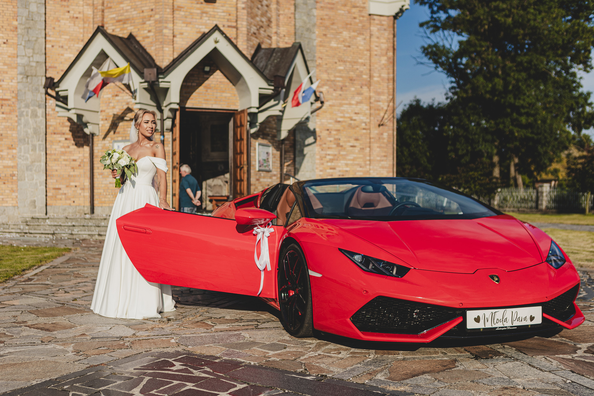 Panna młoda w białej sukni z bukietem kwiatów stoi obok czerwonego Lamborghini z otwartymi drzwiami, na tle kościoła z biało-czerwonymi flagami.