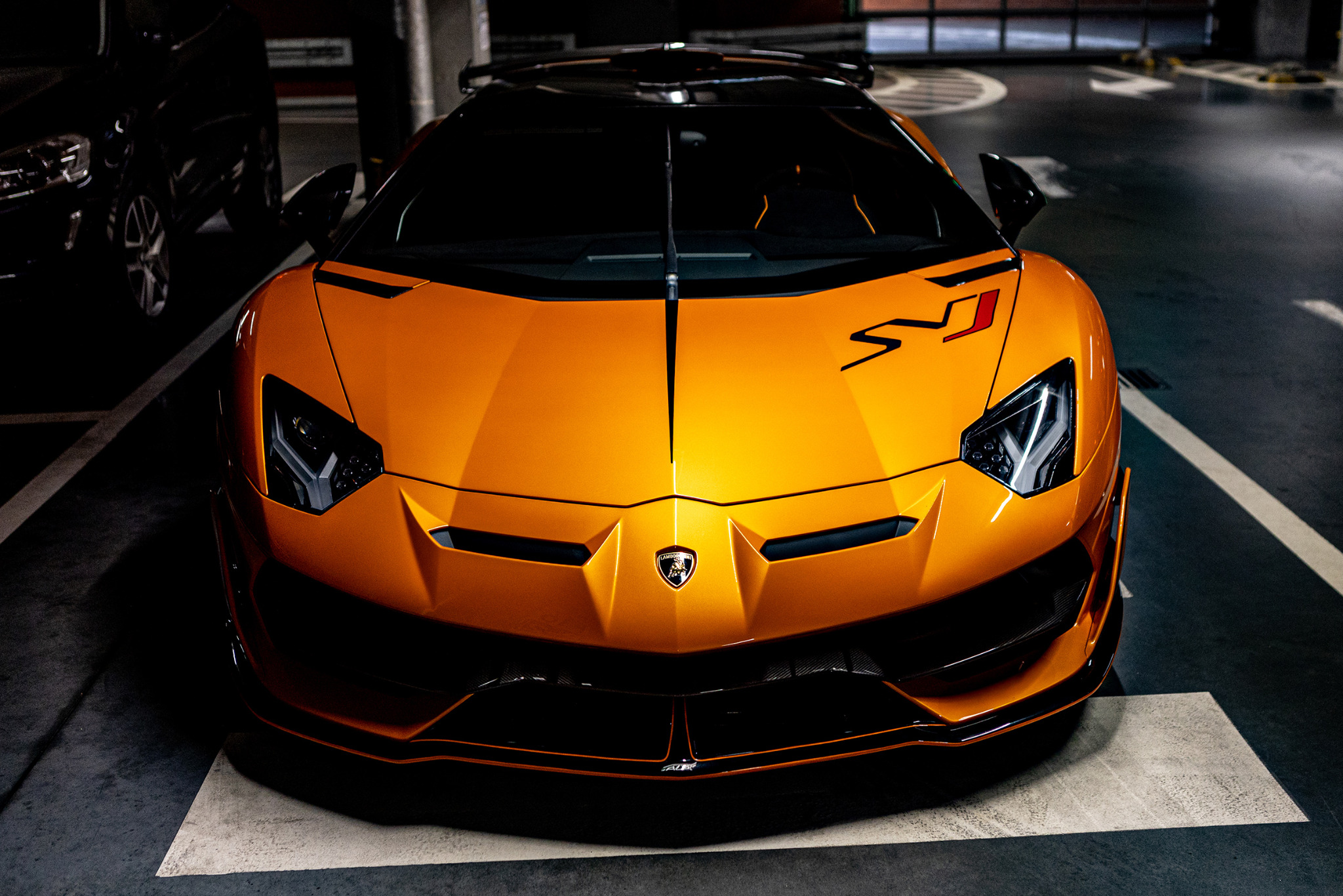 Pomarańczowy Lamborghini Aventador SVJ, widok z przodu, z czarnymi akcentami i naklejkami, zaparkowany w garażu podziemnym.