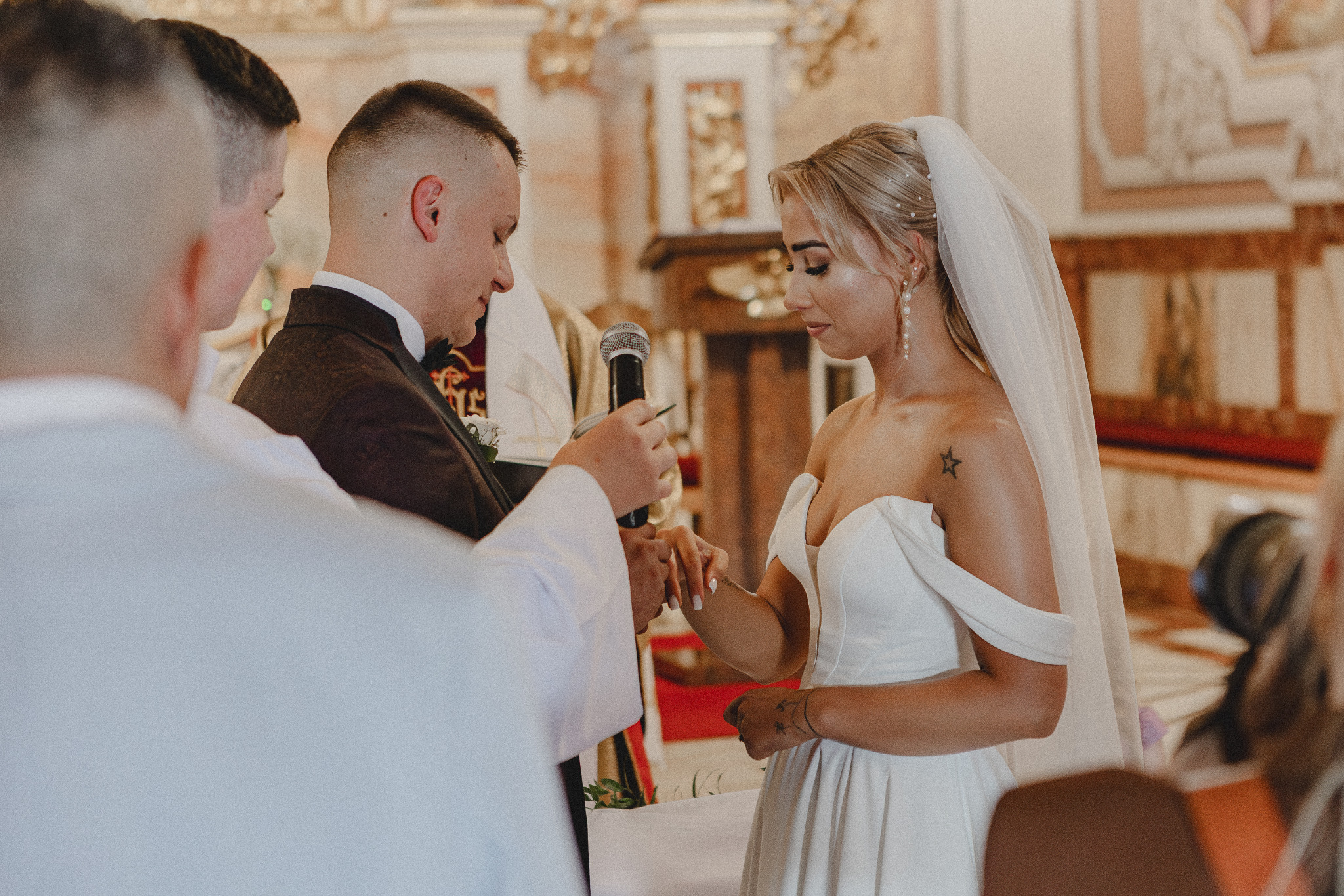 Para młoda podczas ceremonii ślubnej w kościele, wymiana obrączek, panna młoda w białej sukni z odkrytymi ramionami i welonem, pan młody w ciemnym garniturze, ksiądz trzyma mikrofon.