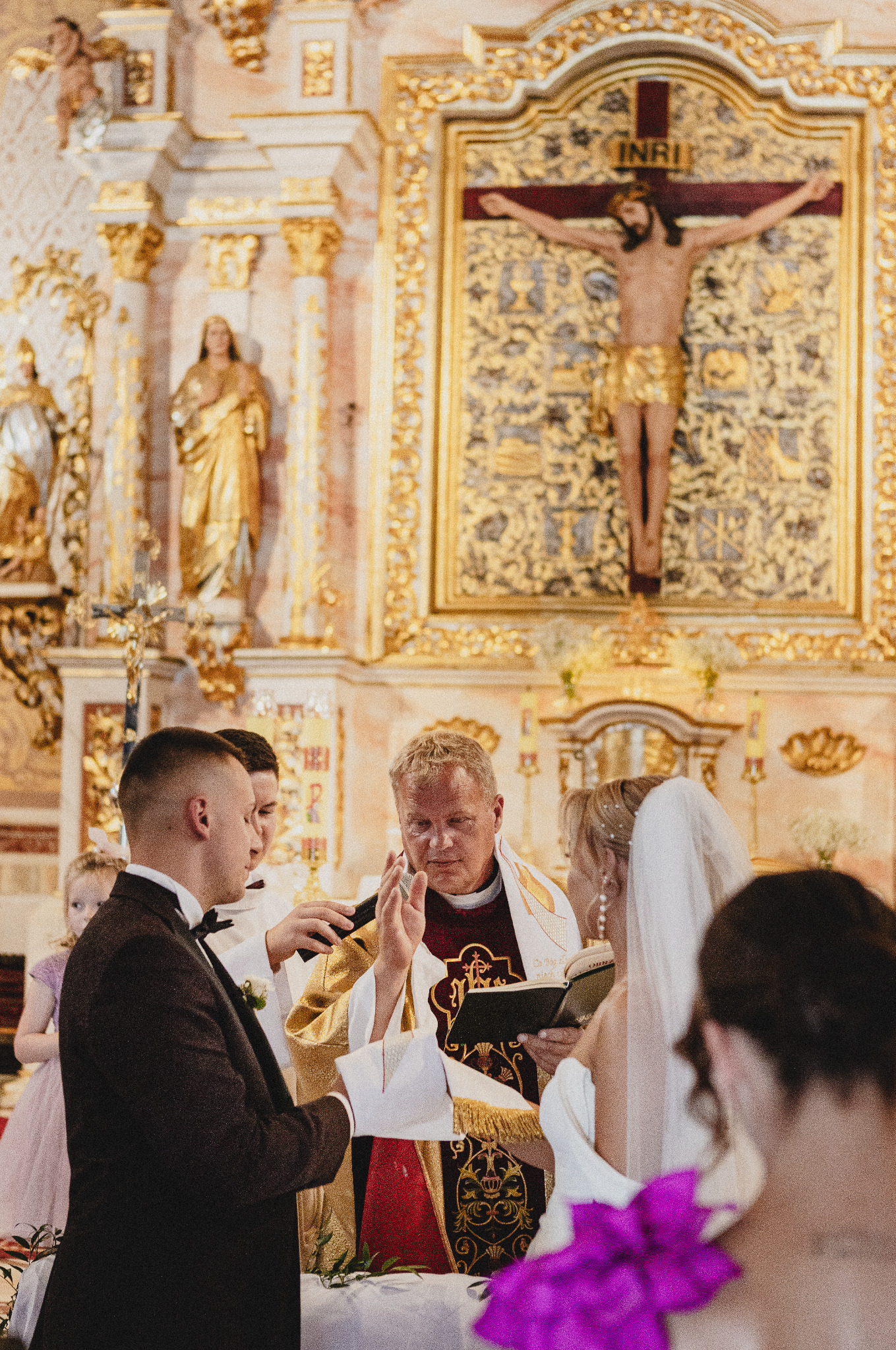 Uroczysta ceremonia ślubna w bogato zdobionym kościele; ksiądz błogosławi parę młodą podczas czytania z księgi, na tle złoconego ołtarza z krucyfiksem.
