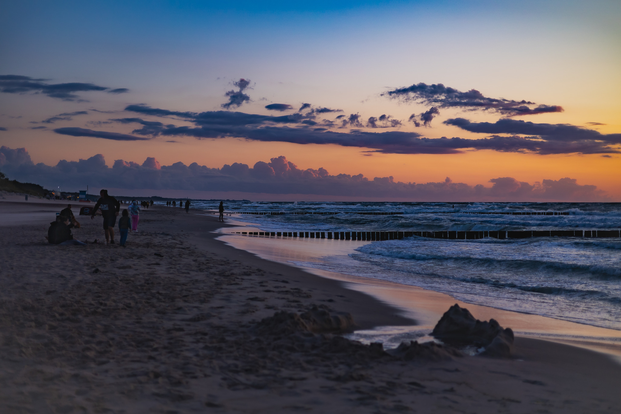 Plaża o zmierzchu z widocznymi falami uderzającymi o drewniane falochrony, sylwetki ludzi spacerujących w oddali, niebo w odcieniach pomarańczy i błękitu.