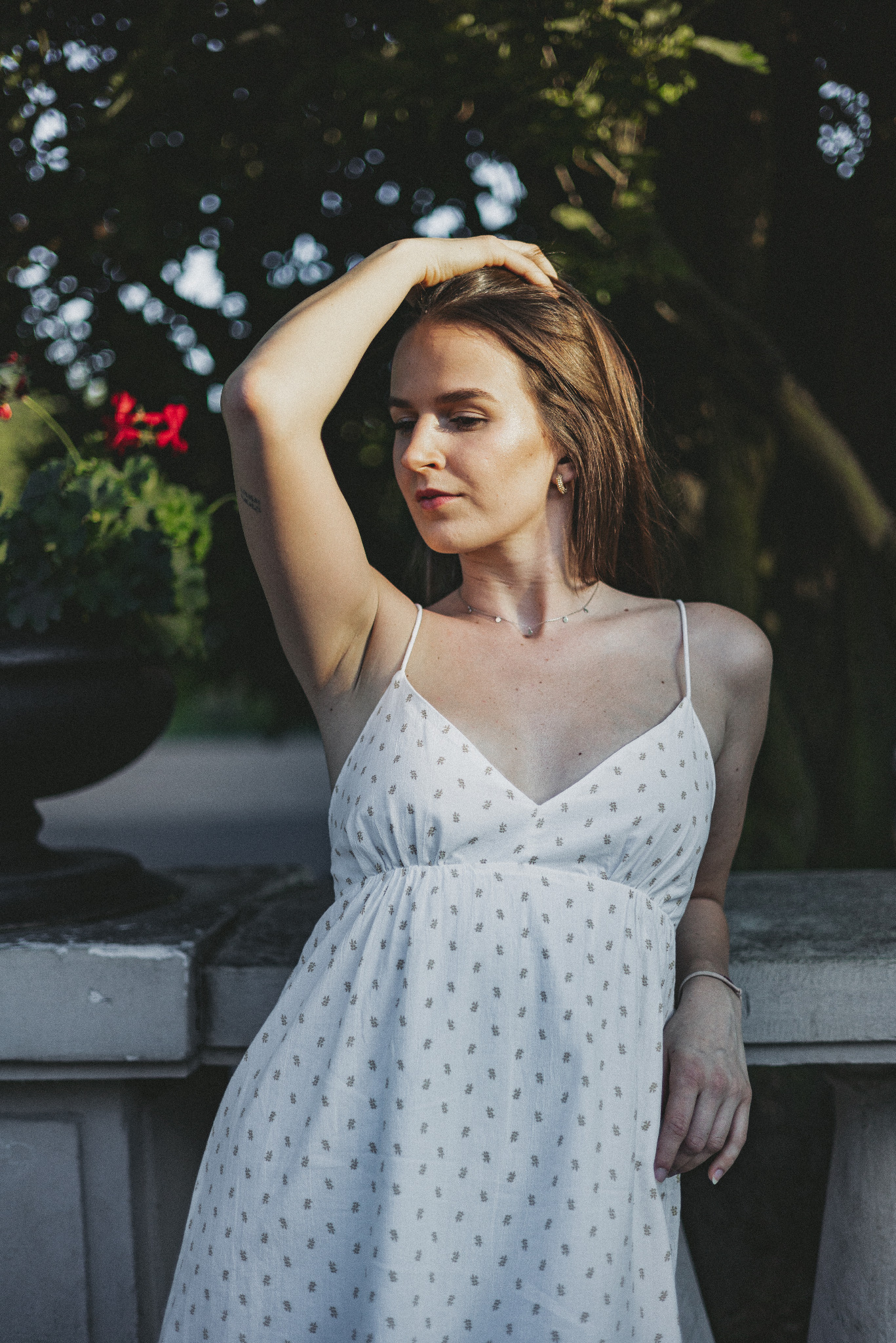 Portret kobiety w letniej sukience, opierającej się o kamienną balustradę w parku, z ręką uniesioną do włosów i delikatnym naszyjnikiem.