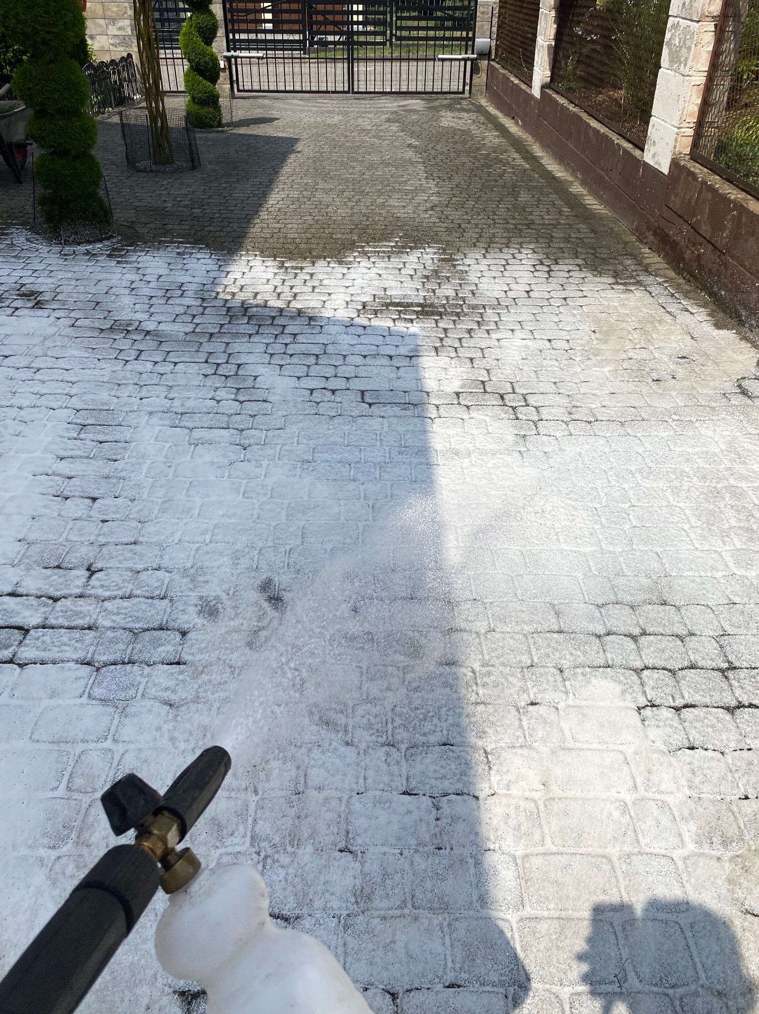 Częściowo wyczyszczona kostka brukowa z pianą, widoczna dysza myjki ciśnieniowej, brama i zadbany ogród w tle.