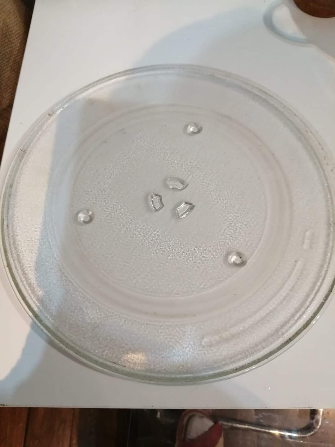 Szklany talerz obrotowy z kuchenki mikrofalowej z trzema odłamanymi fragmentami szkła na środku, leżący na białym blacie.