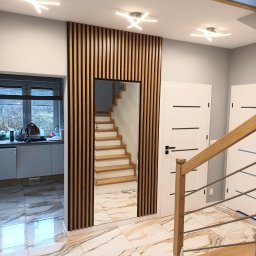 Maciej Szczyrbak MMEBLE MEBLE NA WYMIAR - Nowoczesny przedpokój z lustrem otoczonym drewnianymi lamelami, widok na schody i fragment zabudowy kuchennej z oknem.