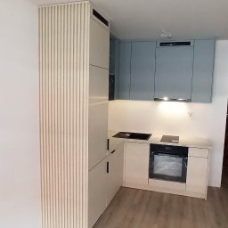 Maciej Szczyrbak MMEBLE MEBLE NA WYMIAR - Jasna kuchnia w kształcie litery L z frontami w kolorze beżowym i błękitnym, z pionowymi żłobieniami na wysokiej szafce, płyta indukcyjna i piekarnik w zabudowie, oświetlenie podszafkowe.