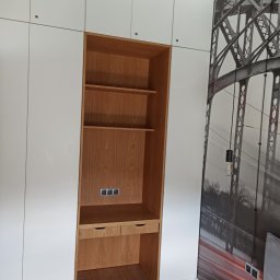 Maciej Szczyrbak MMEBLE MEBLE NA WYMIAR - Biała zabudowa meblowa z drewnianym regałem z półkami i biurkiem, widoczne gniazdka elektryczne, na ścianie fototapeta z motywem mostu.