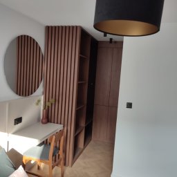 Maciej Szczyrbak MMEBLE MEBLE NA WYMIAR - Nowoczesna szafa wnękowa z drewnianymi lamelami i otwartymi półkami, zintegrowana z biurkiem i okrągłym lustrem w minimalistycznym wnętrzu sypialni, oświetlona wiszącą lampą z czarnym abażurem...