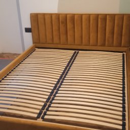 Maciej Szczyrbak MMEBLE MEBLE NA WYMIAR - Rama łóżka z tapicerowanym, musztardowym zagłówkiem w pionowe pasy i drewnianym stelażem listwowym, widok z przodu na tle ściany w trakcie malowania.