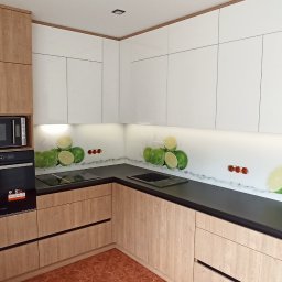 Maciej Szczyrbak MMEBLE MEBLE NA WYMIAR - Nowoczesna kuchnia z frontami w kolorze jasnego drewna i białymi szafkami górnymi, czarnym blatem i panelem szklanym z motywem limonek. W zabudowie czarna lodówka Samsung z odbijającym się fotografem.