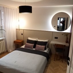 Maciej Szczyrbak MMEBLE MEBLE NA WYMIAR - Nowoczesna sypialnia z łóżkiem, drewnianą podłogą ułożoną w jodełkę, okrągłym lustrem z podświetleniem LED i biurkiem z krzesłem.