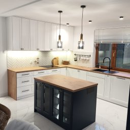 Maciej Szczyrbak MMEBLE MEBLE NA WYMIAR - Nowoczesna kuchnia z białą zabudową, drewnianym blatem i czarną wyspą z przeszklonymi szafkami, oświetlona wiszącymi lampami z elementami mosiądzu.